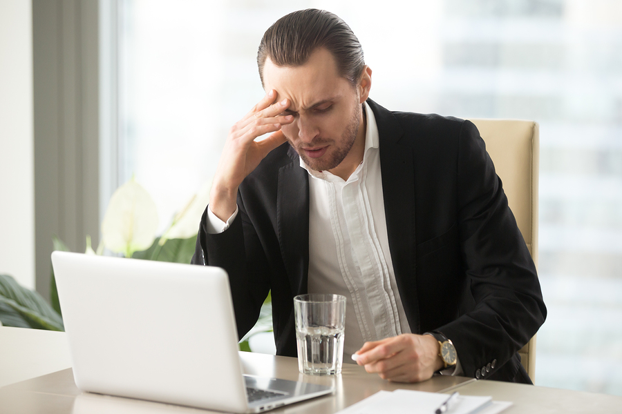 Stressed businessman having severe headache, holding painkiller remedy pill. Sick CEO with head cold, thinking about taking medicine with glass of water. Being ill at work, stress induced migraine.