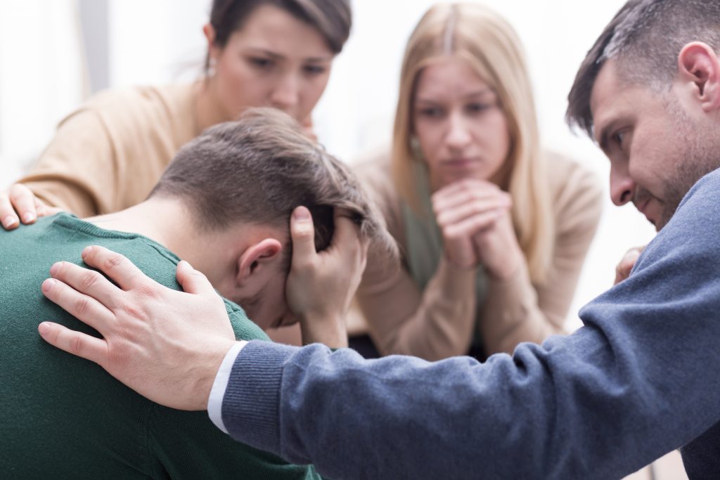 Close-up of a devastated young man holding his head in his hands and a group of friends in a supportive pose around him