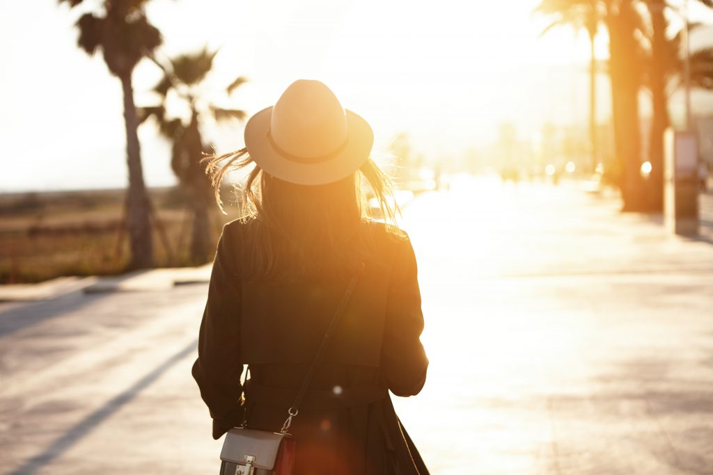 Back view of slender young woman wearing hat and coat walking down street on sunny spring day heading to city beach to enjoy beautiful sunset. People lifestyle travel and vacations concept