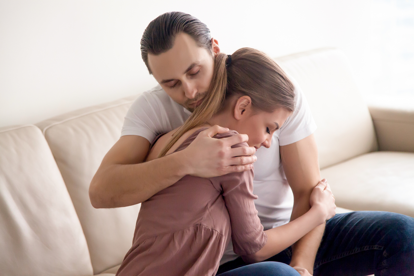 bigstock–185095969 (1) Young tender husband hugging his wife, loving man embracing woman gently and tightly sitting on couch at home, showing love and support, happy soulmates or close friends, peace, harmony, understanding