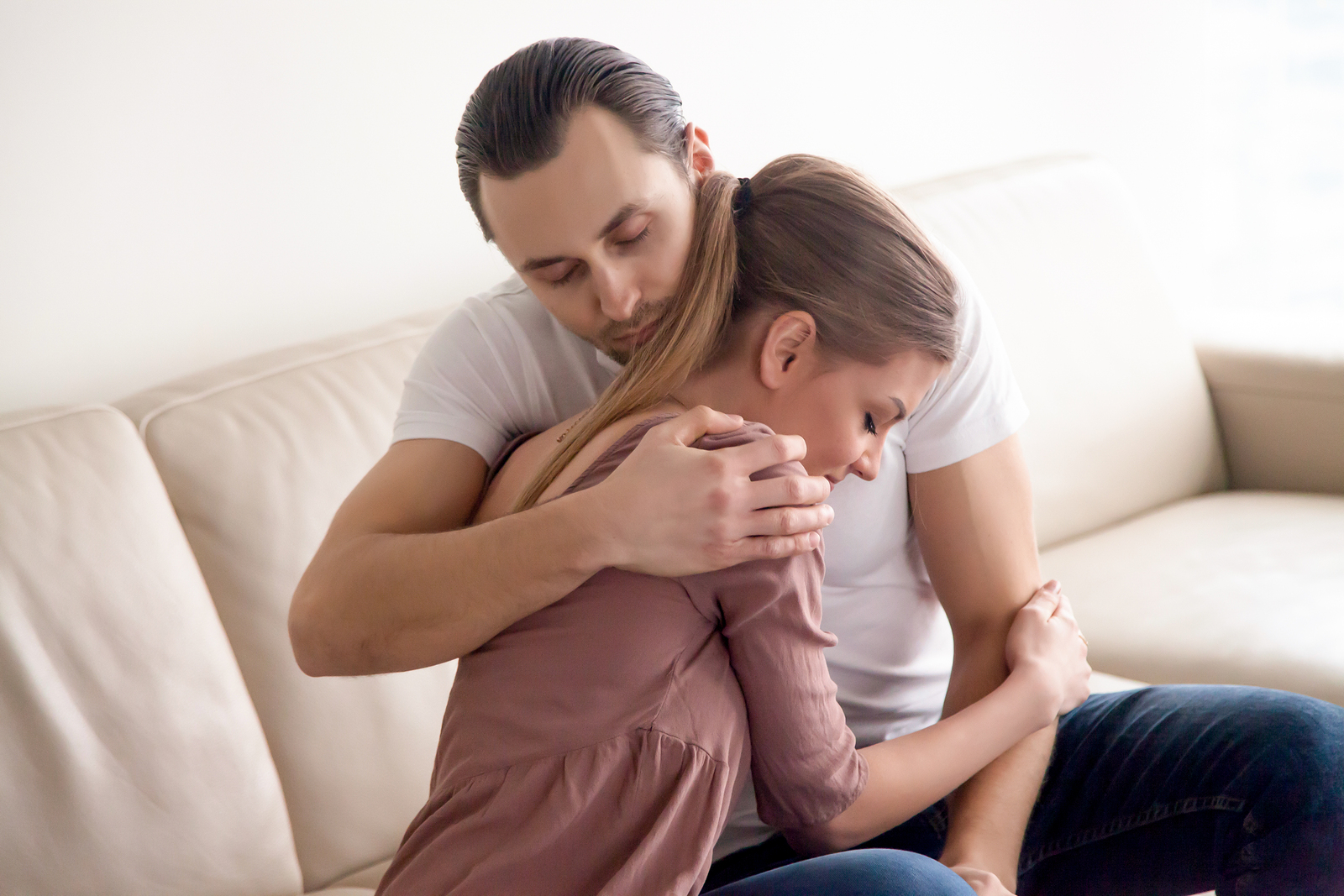 Young tender husband hugging his wife, loving man embracing woman gently and tightly sitting on couch at home, showing love and support, happy soulmates or close friends, peace, harmony, understanding