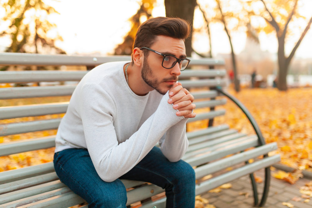 A lonely handsome man sits sad in the park on a bench. Autumn season, yellow leaves on background. Loneliness concept. The student did not pass the exams or broke up with the girlfriend