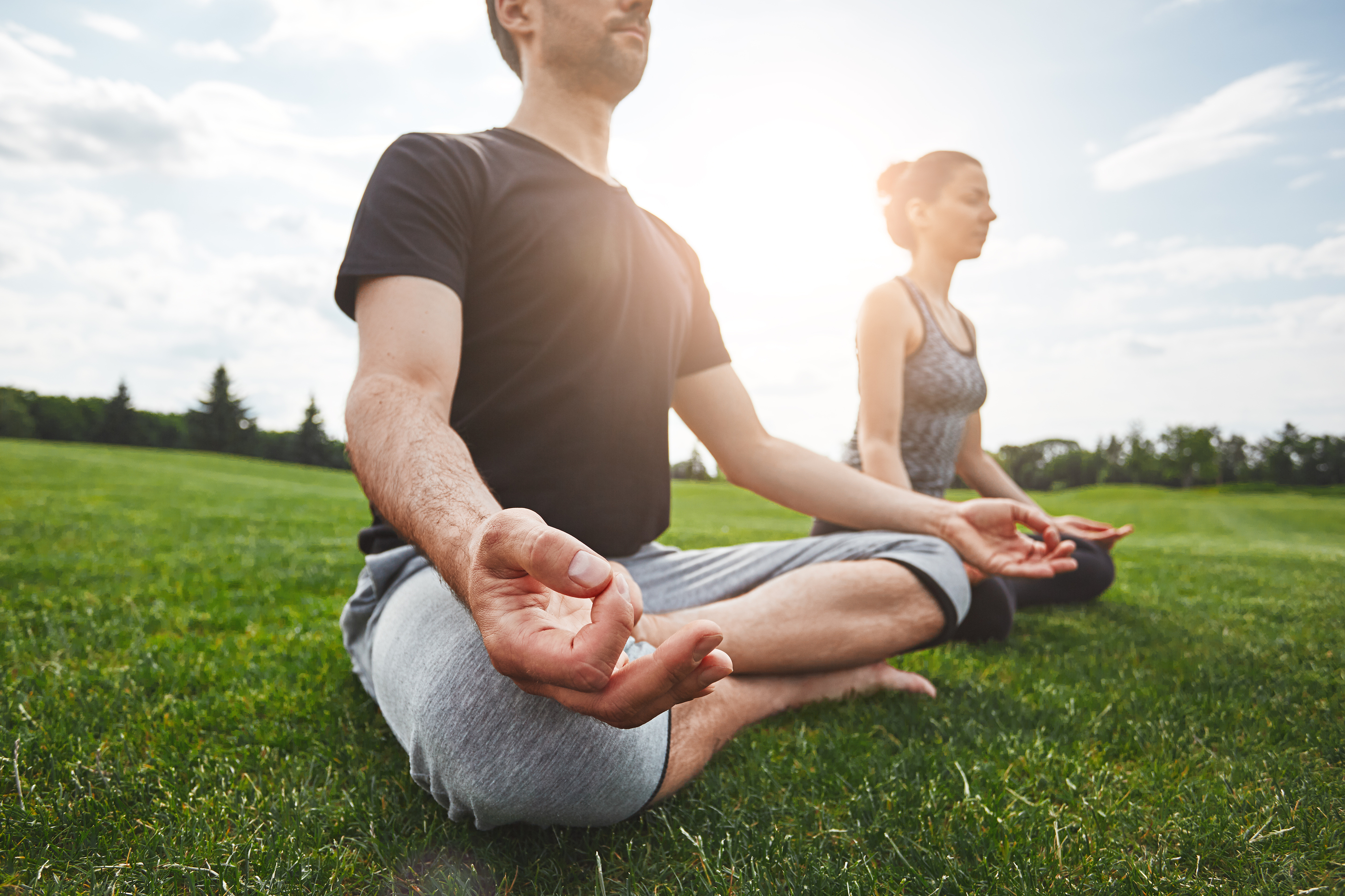 Yoga class. Young couple are meditating and doing yoga exercises while sitting in lotus pose on a green grass in open field. Sunny morning. Meditation concept