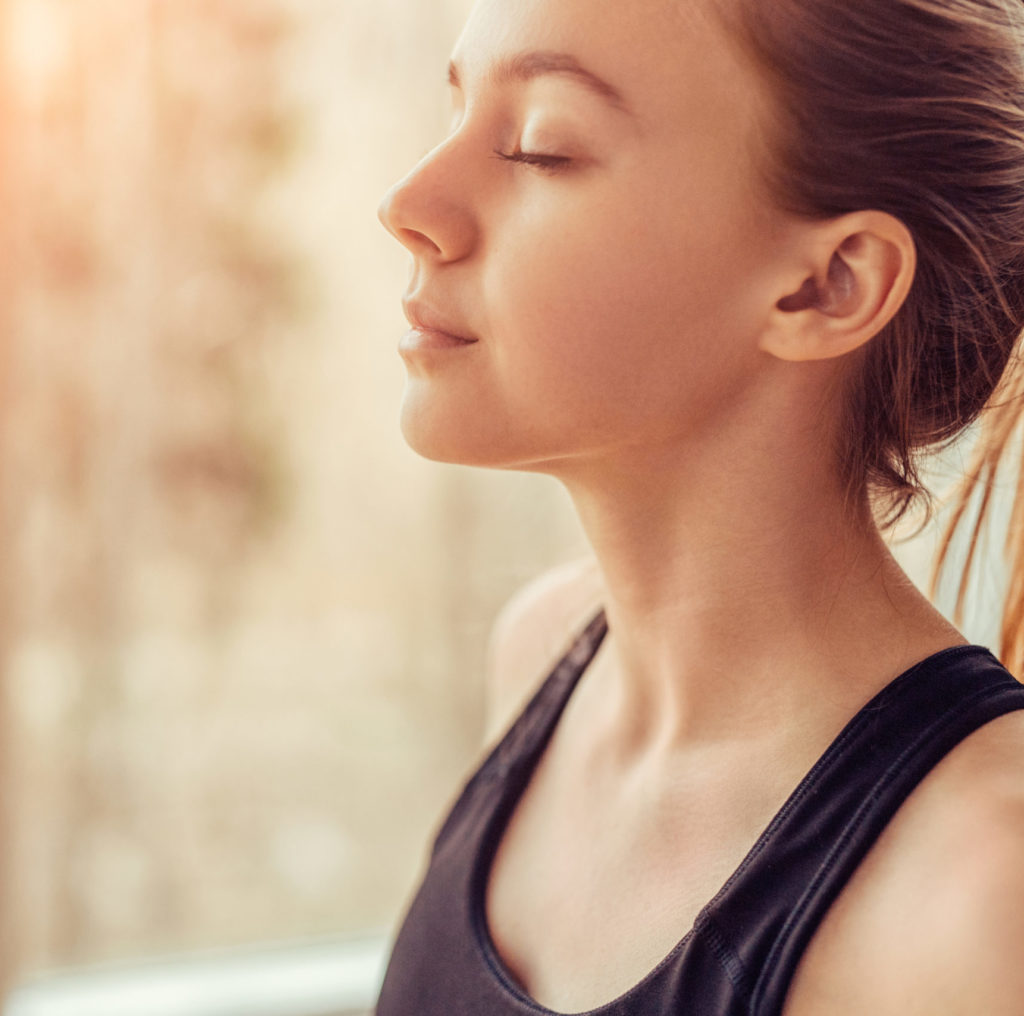 Side view of young female with closed eyes breathing deeply while doing respiration exercise during yoga session in gym