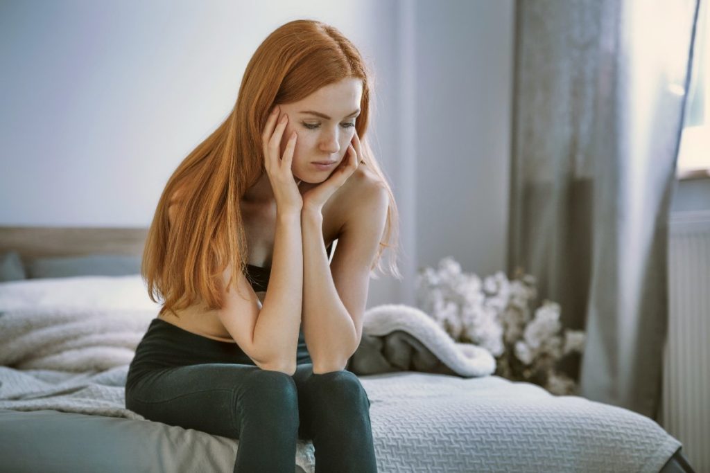 A teenager sits on the edge of her bed with her head resting in her hands. She is visibly upset and struggling with her body image.