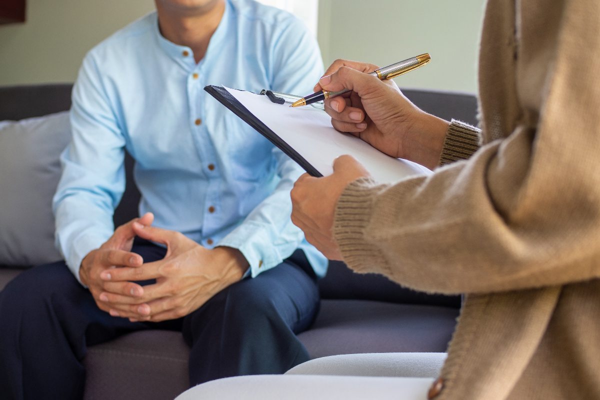 A patient sits across from a therapist during a treatment session for depression. The therapist is holding a clipboard and taking notes, while the patient sits across from the therapist, explaining their symptoms.