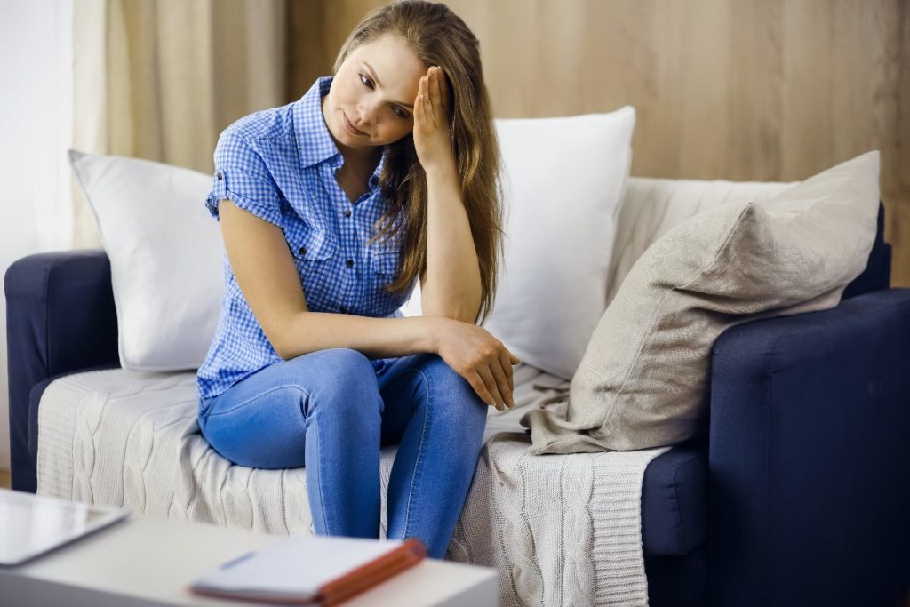 A young woman sits on a couch wit her head resting in her hand. She is visibility upset and struggling to cope with her emotions.