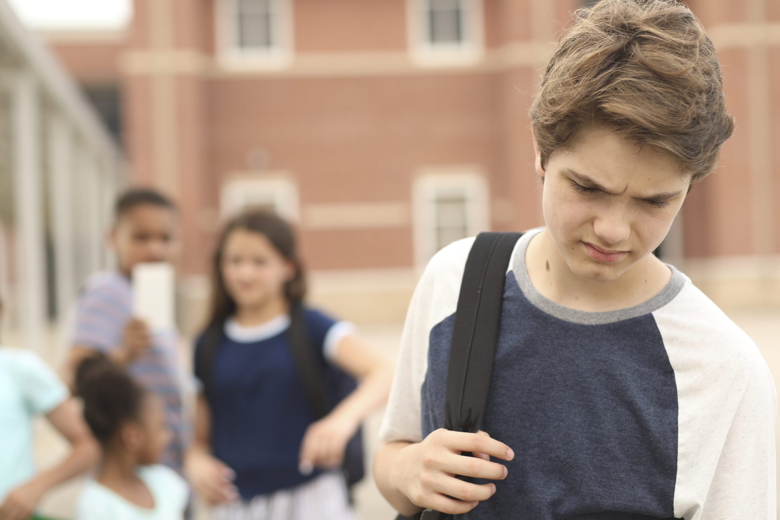 Sad, junior high school age boy being bullied outside the school building, A group of multi-ethnic students in background laugh at the boy. They have mobile devices to cyber bully the child as well as point and talk about him.