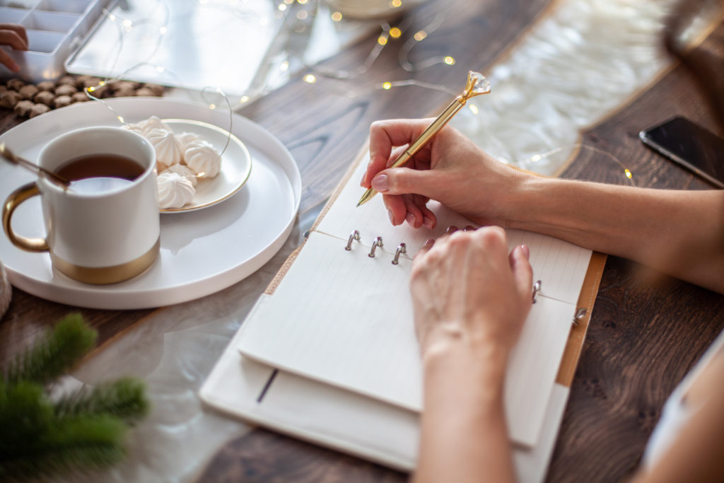 Young woman drinking tea and writing plans or goals for New Year 2021 while her daughter crafting Christmas trees from paper cone, yarns and buttons with placed stars and fairy lights on wooden table.