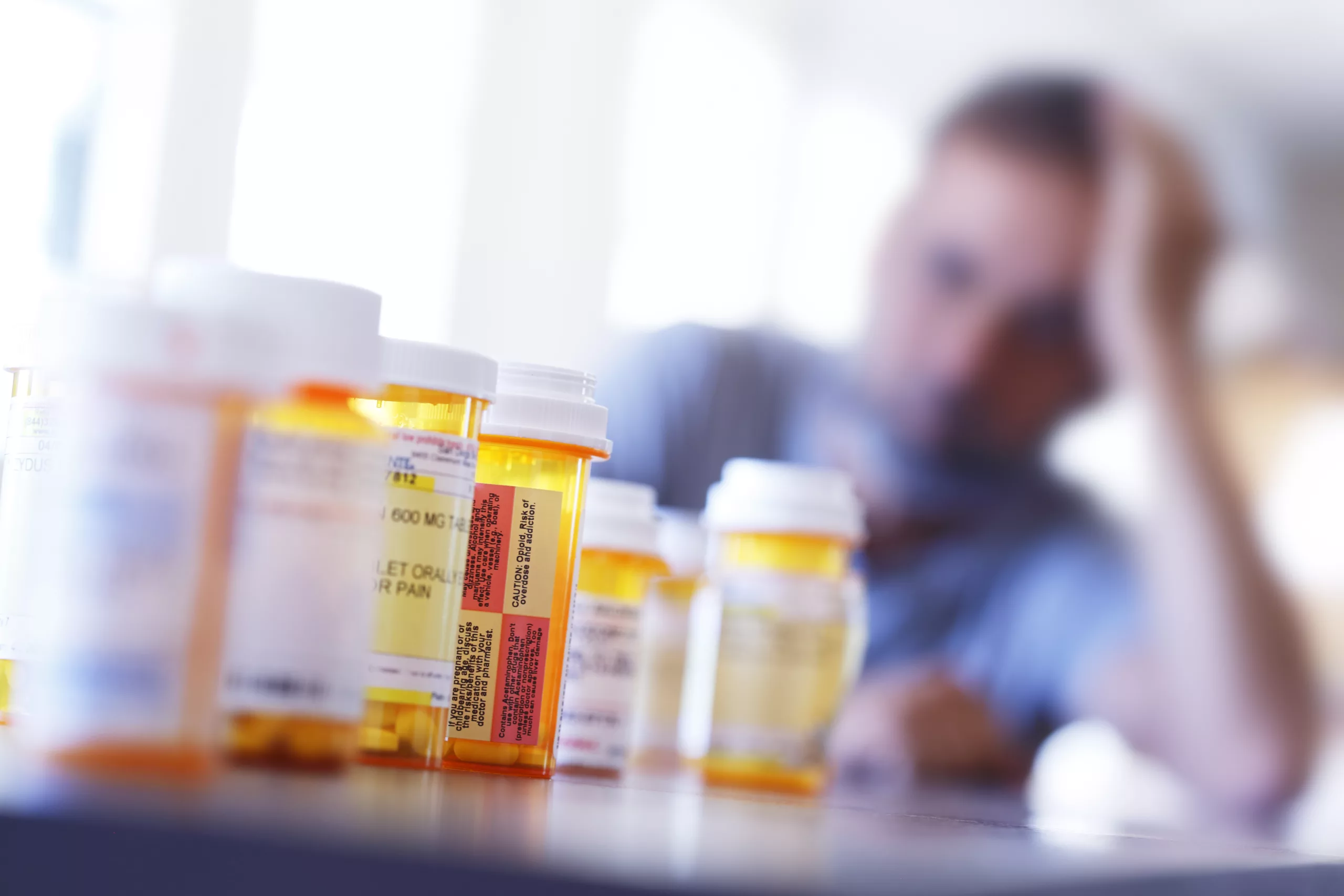 A large group of prescription medication bottles sit on a table in front of a distraught man who is leaning on his hand as he sits at his dining room table. The image is photographed with a very shallow depth of field with the focus being on the pill bottles in the foreground.
