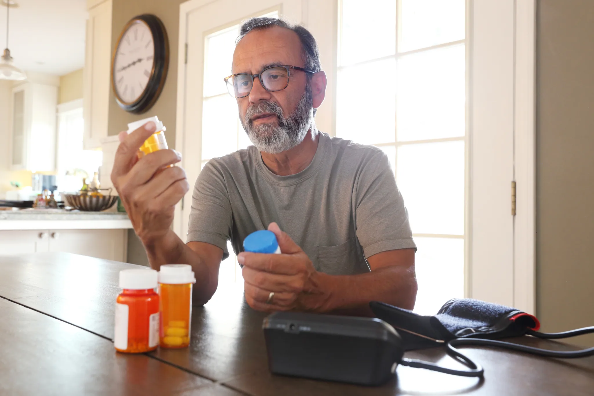A man sits at his dining room table and sorts through various prescription medications as sunlight filters in through the window behind him bathing the room with a soft glow of light. A blood pressure machine rests on the table in front of him.