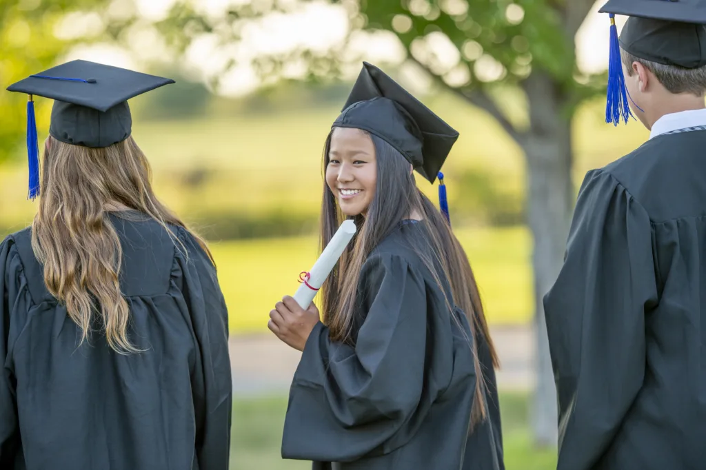 A young female graduate turns around as she poses for a portrait.  She is wearing a gown and cap and holding her diploma as she stands with her peers who have their backs to the camera.