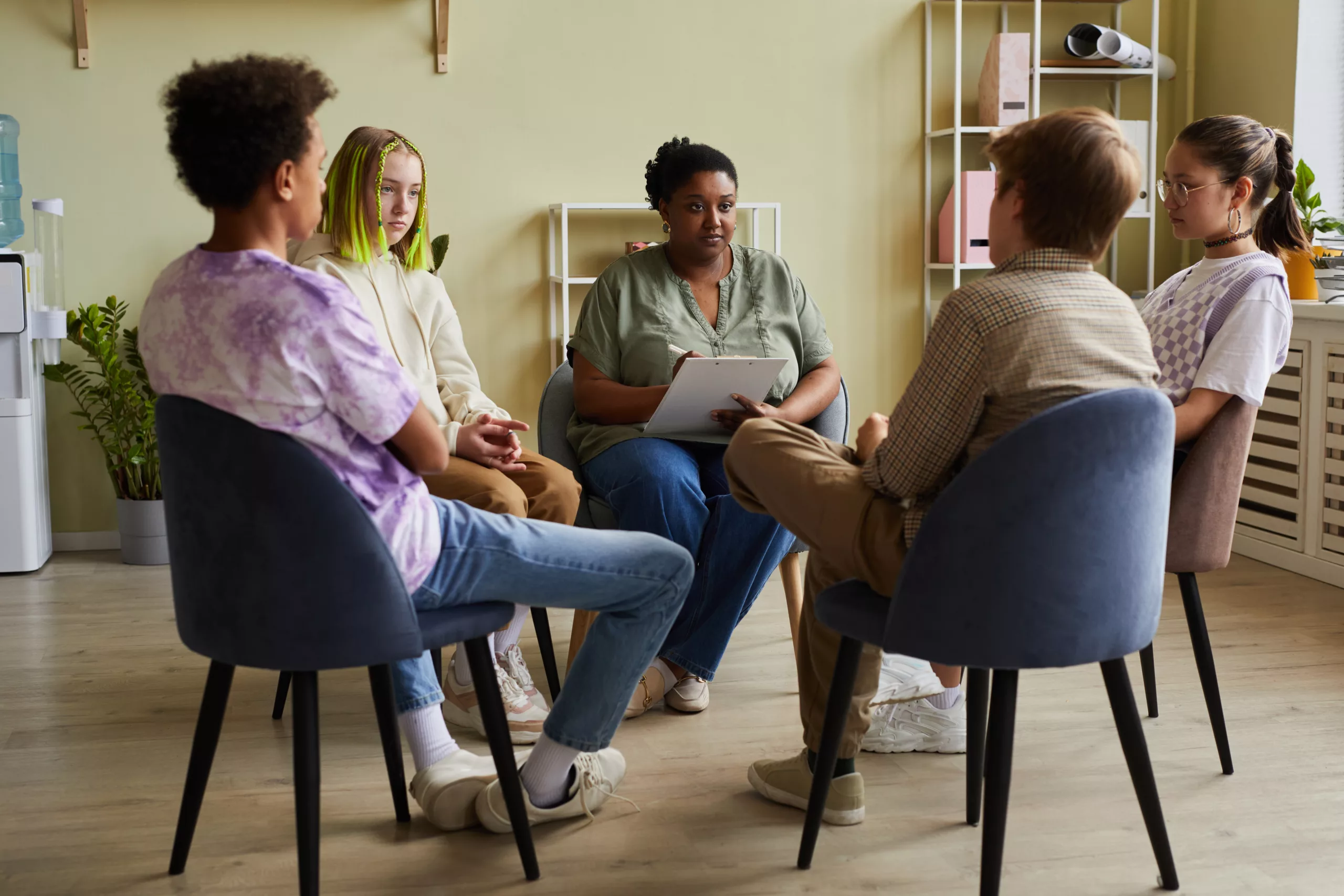 African female psychologist making notes and talking to group of difficult teenagers during therapy session