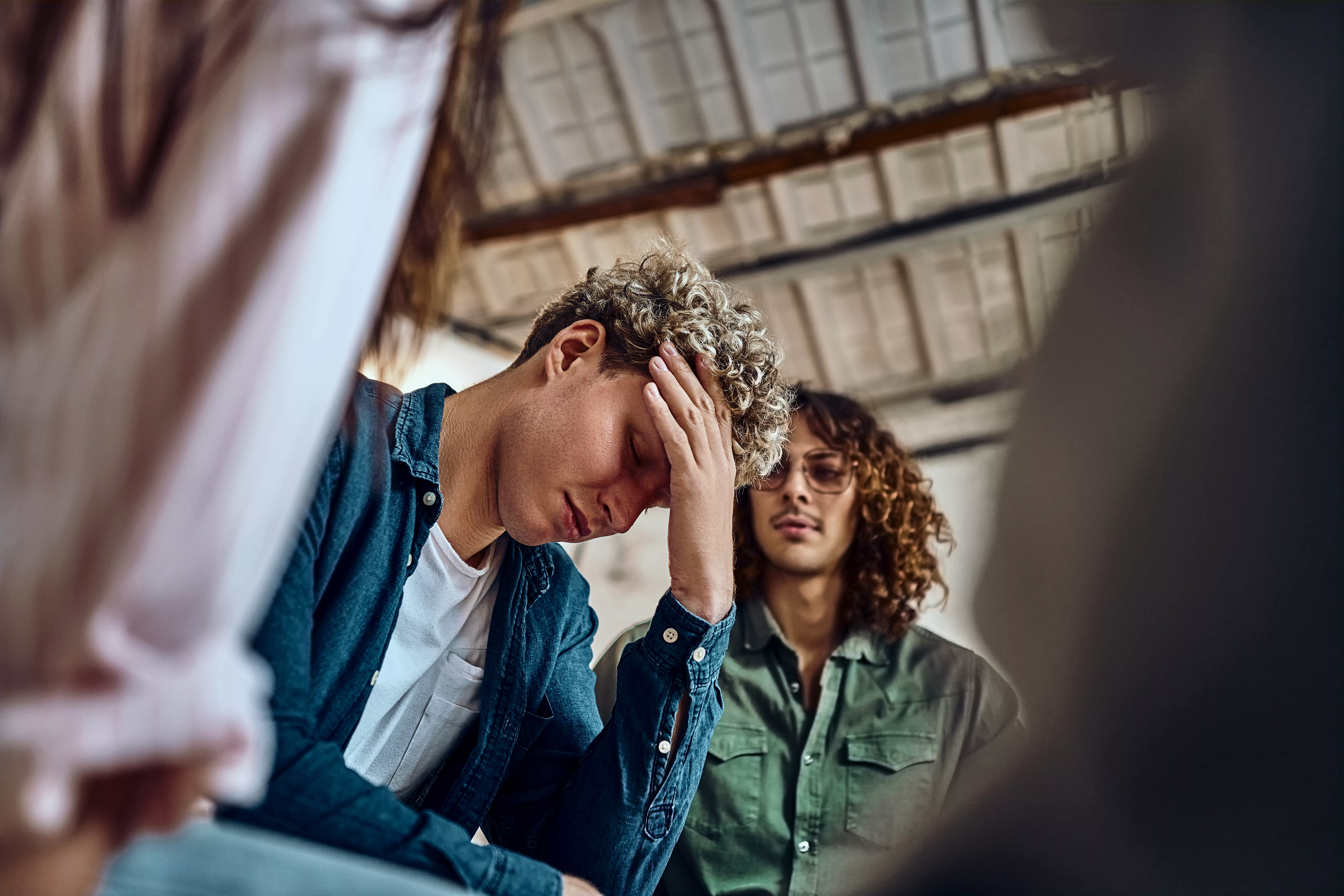 Close-up view of a devastated young man holding his head in his hands and a group of friends in a supportive pose around him. Selective focus