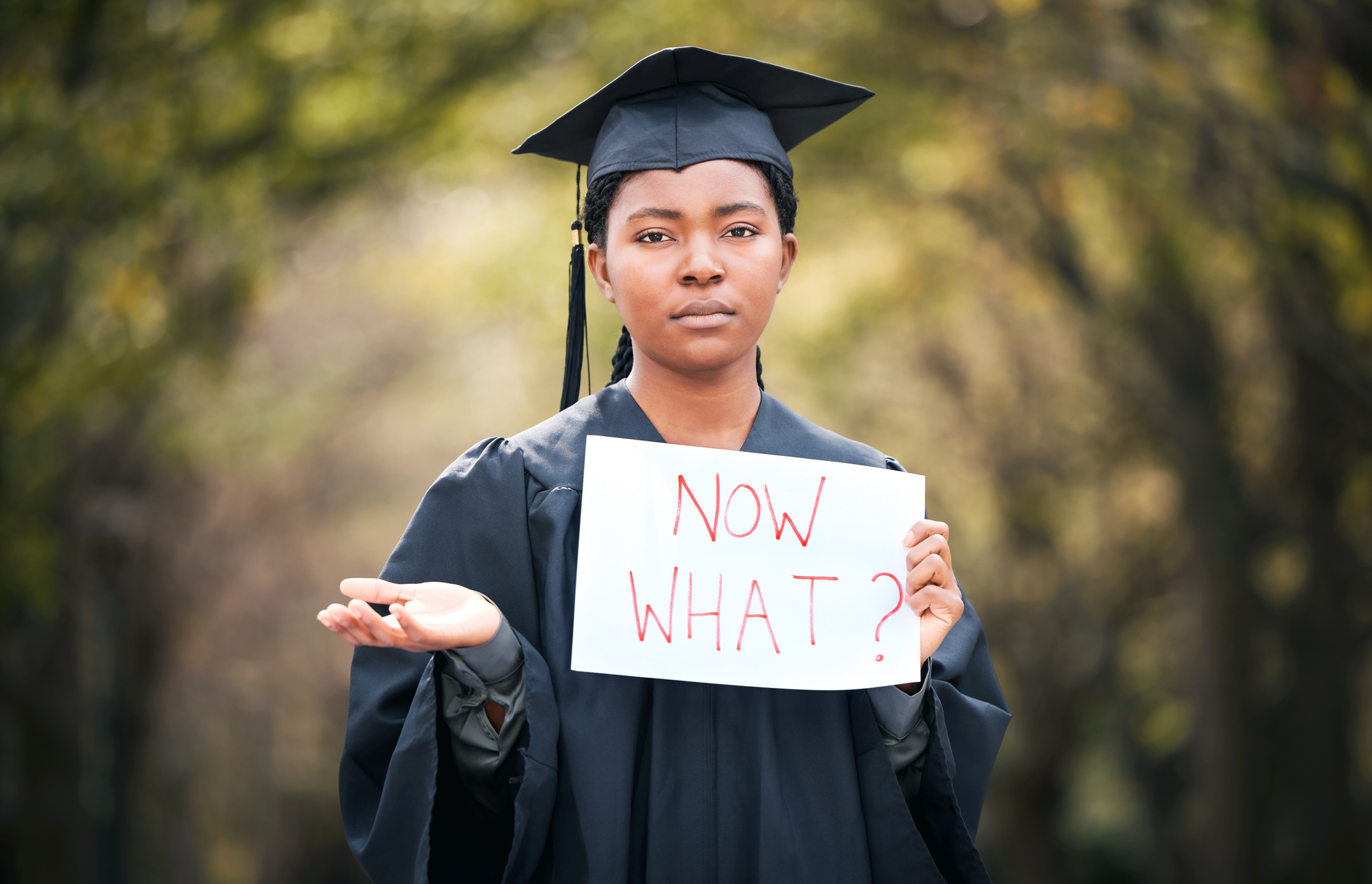 Portrait of a young woman holding a sign that reads "now what" on graduation day