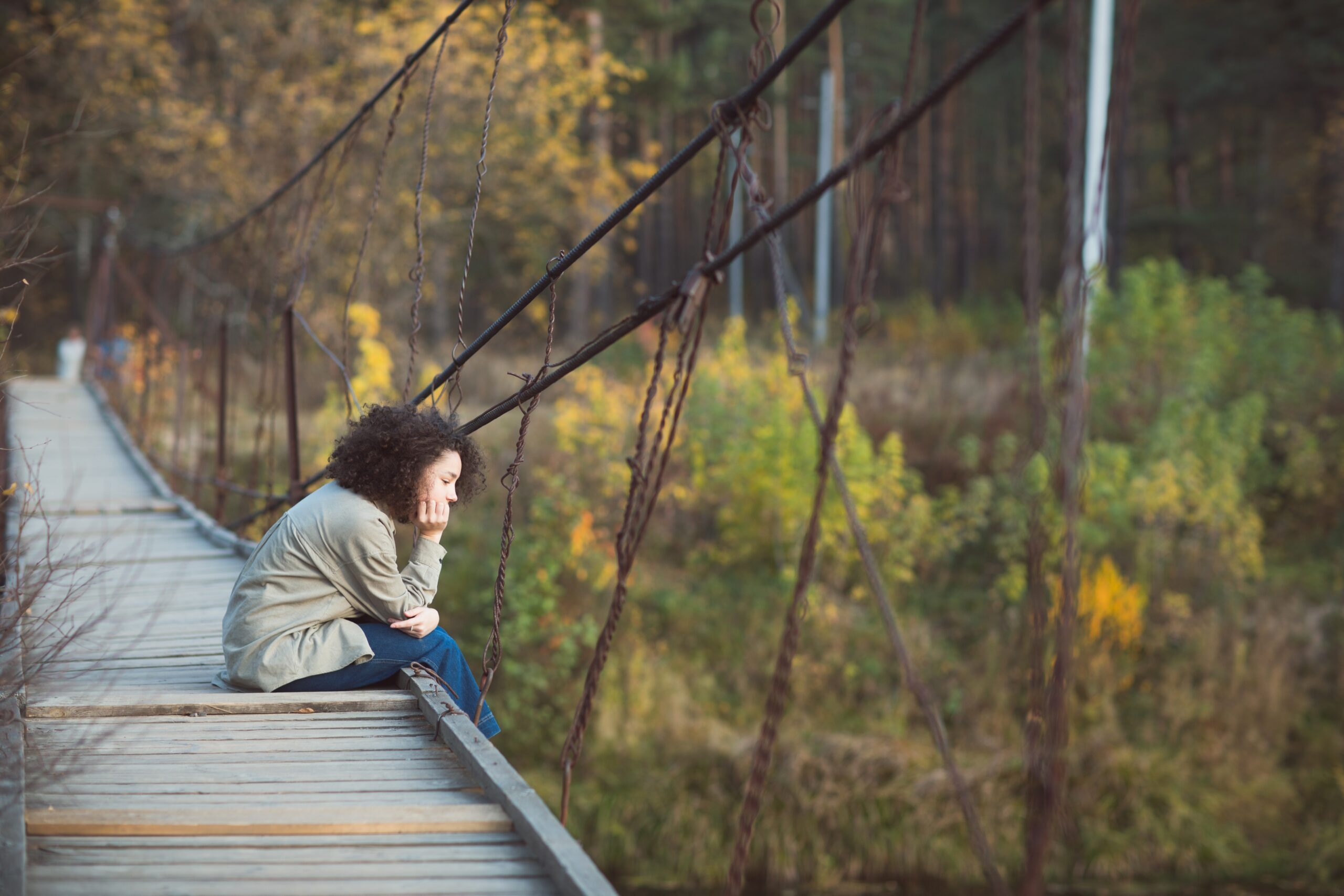 girl with curly hair sits on bridge over river in countryside in autumn. outdoor recreation alone.