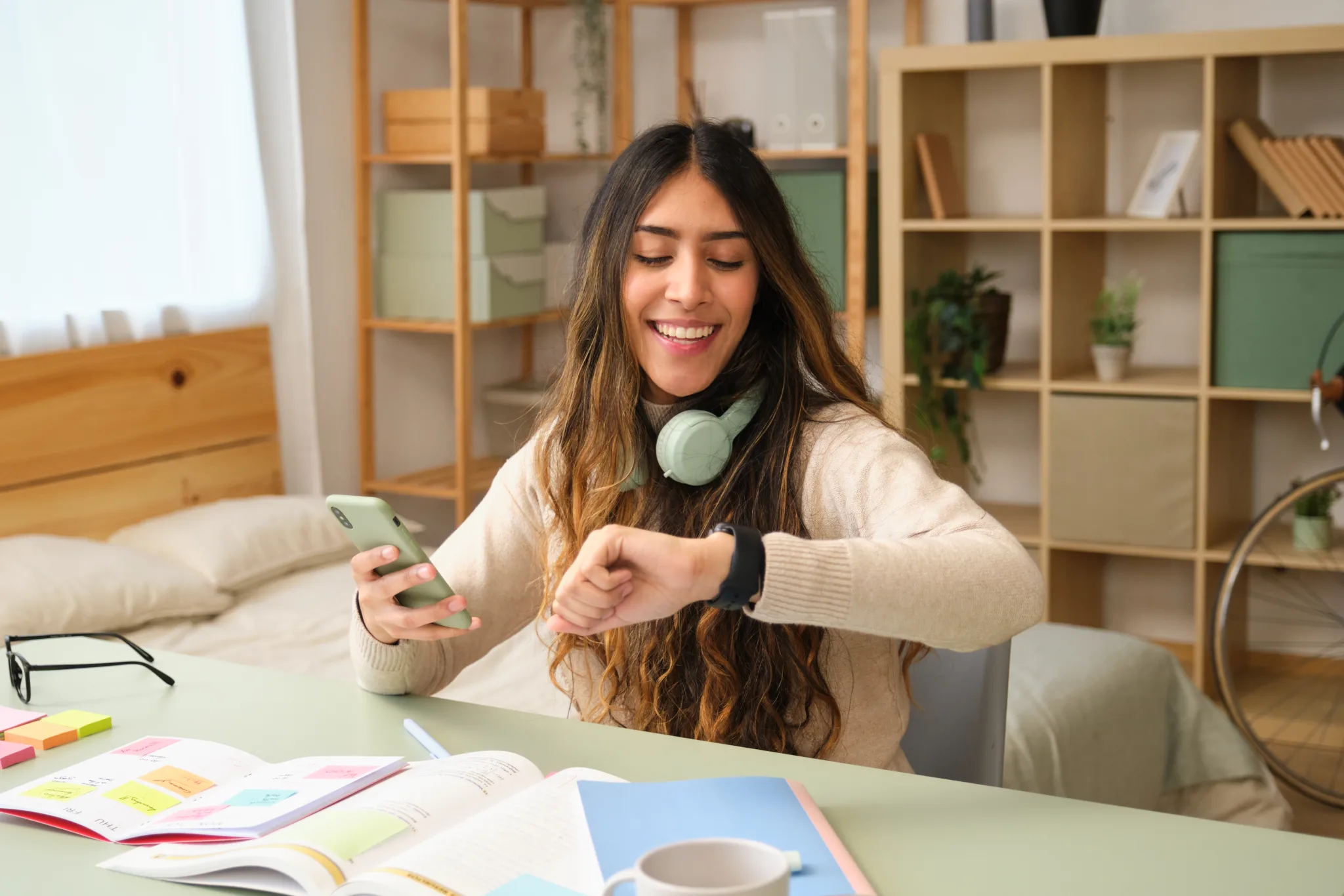 Happy student checking the time on her smartwatch while holding her phone and studying at her desk