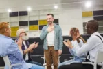 Portrait of smiling mature man  introducing himself during therapy session in support group to people clapping, copy space