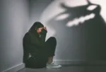 Shot of a young woman sitting in the corner of a dark room with a scary figure on the wall.