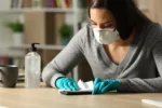 Woman with gloves and protective mask disinfecting smart phone from coronavirus sitting on a desk at night at home