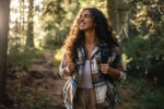 Young woman with curly hair, wearing a backpack, smiles cheerfully while walking through a forest path, feeling the beauty, freedom, and tranquility of nature.