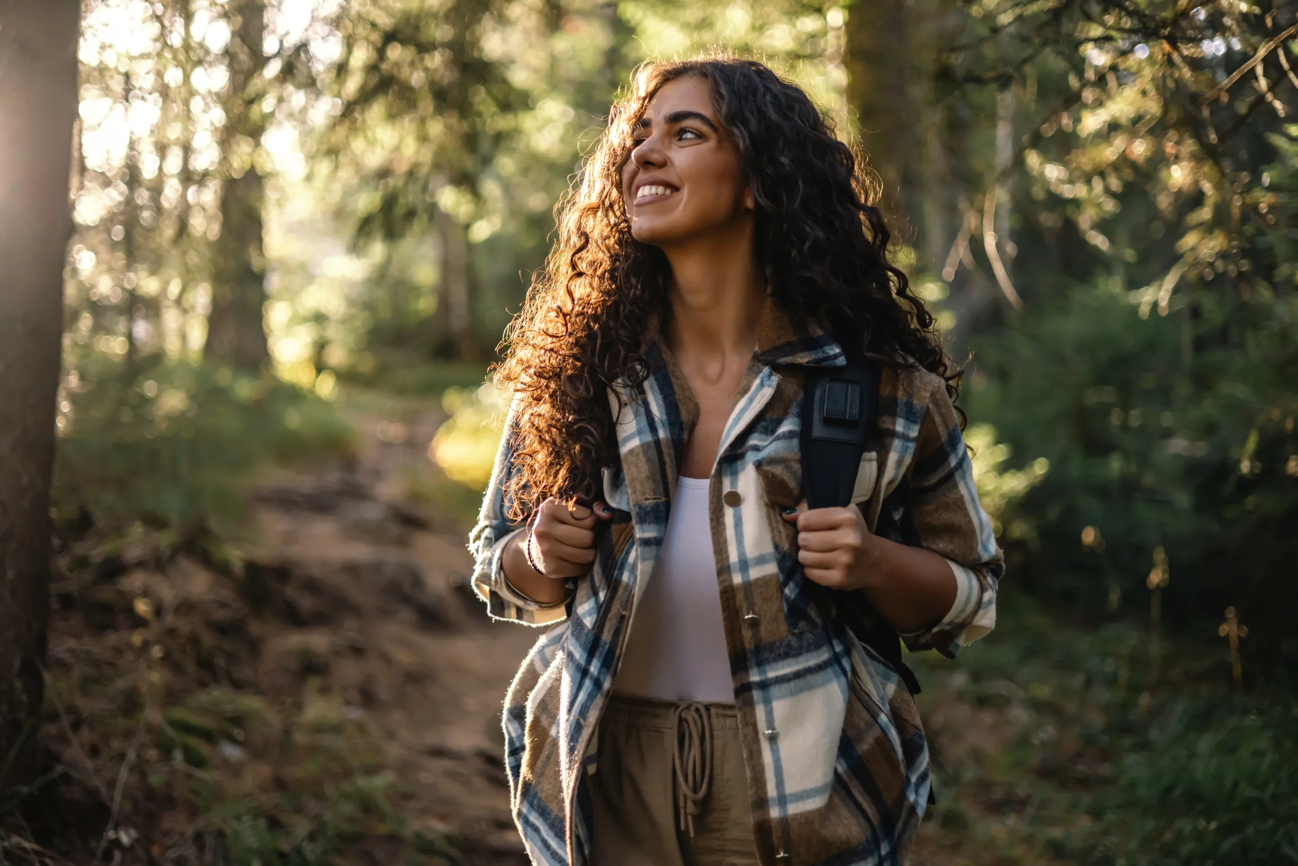 Young woman with curly hair, wearing a backpack, smiles cheerfully while walking through a forest path, feeling the beauty, freedom, and tranquility of nature.